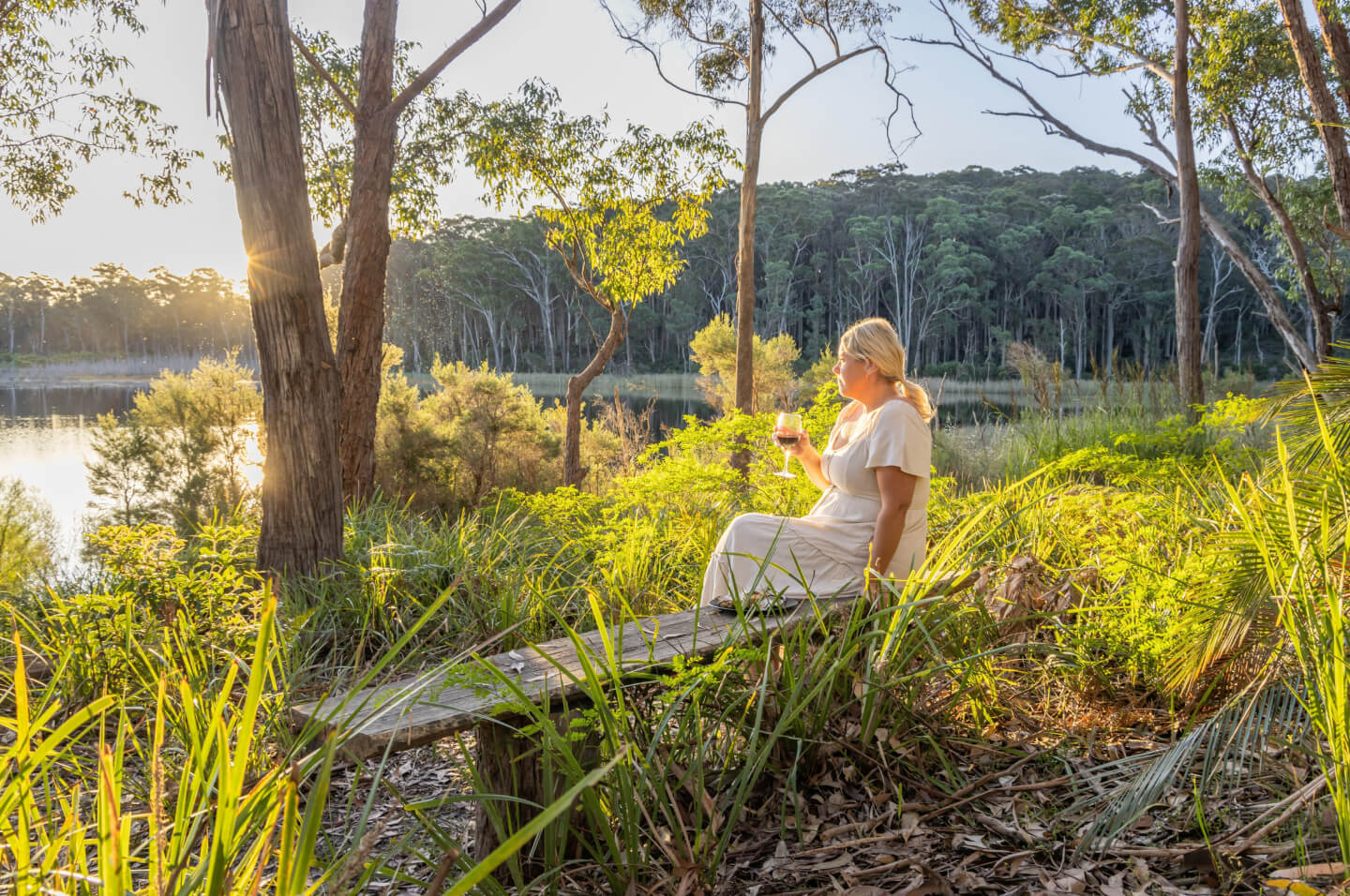A woman in a white dress sits on a rustic wooden bench in a lush natural setting, holding a glass of wine. The scene is bathed in warm golden sunlight, likely during late afternoon or early evening. She is surrounded by tall grasses and trees, with a calm lake and dense forest in the background. The atmosphere is peaceful and serene.